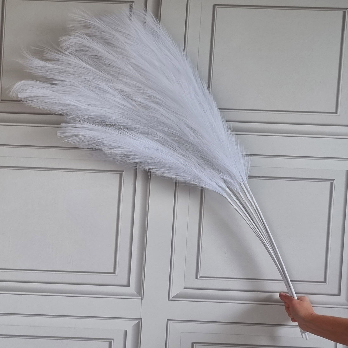 A large white pampas grass decorative stem held by a hand in front of a light gray paneled door.