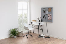 Load image into Gallery viewer, A modern home office setup featuring a white desk with black metal legs, a white swivel chair, a black floor lamp, and a gray corkboard on the wall, set in a bright room with light wood floors and a window.
