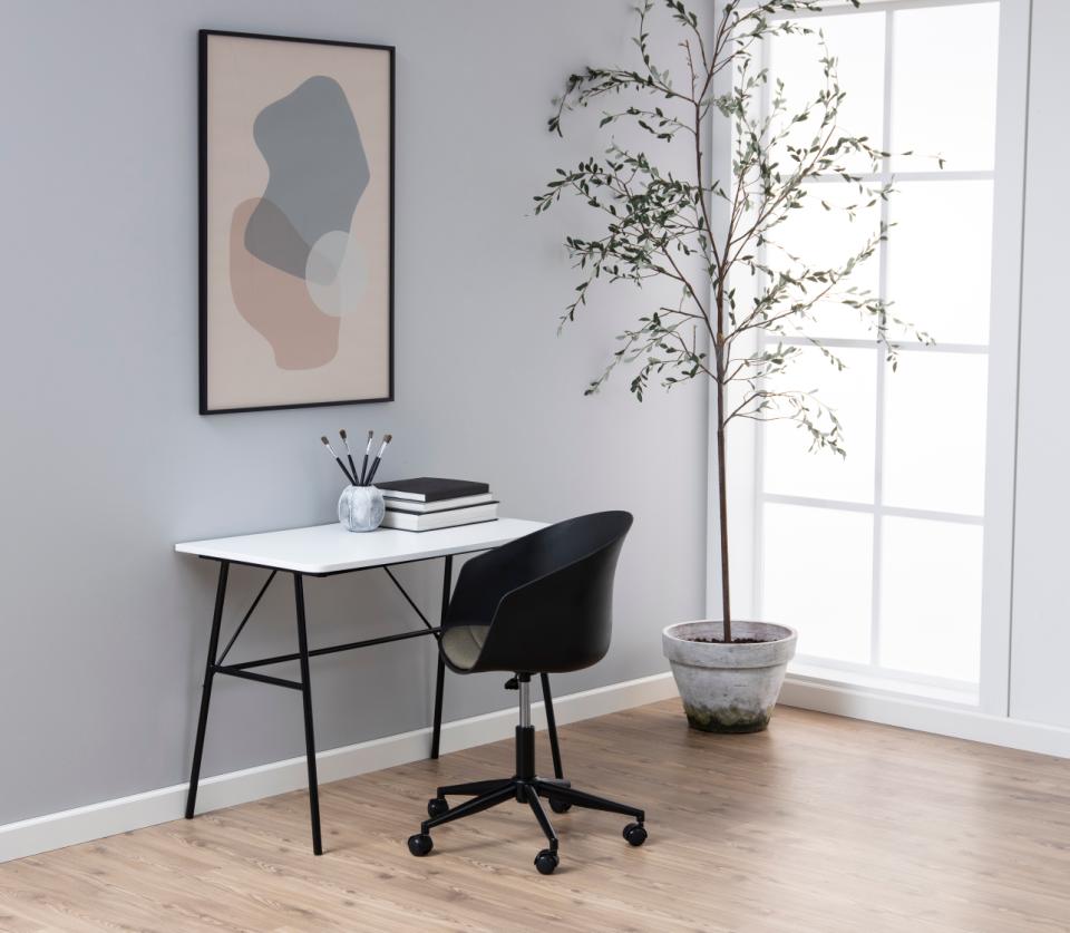 A modern home office setup featuring a white desk with black metal legs, a black swivel office chair, a framed abstract art print on a light gray wall, and a tall potted tree next to a bright window.