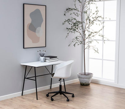 A modern home office setup featuring a white desk with black metal legs, a white swivel office chair, a framed abstract art print on a light gray wall, and a tall potted tree next to a bright window.