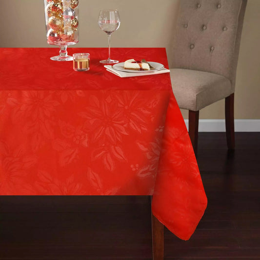 A red Christmas tablecloth with a subtle poinsettia floral pattern is laid out on a rectangular dining table. A plate and glass are set on top of the tablecloth, and there is a chair with a cushion in the background.