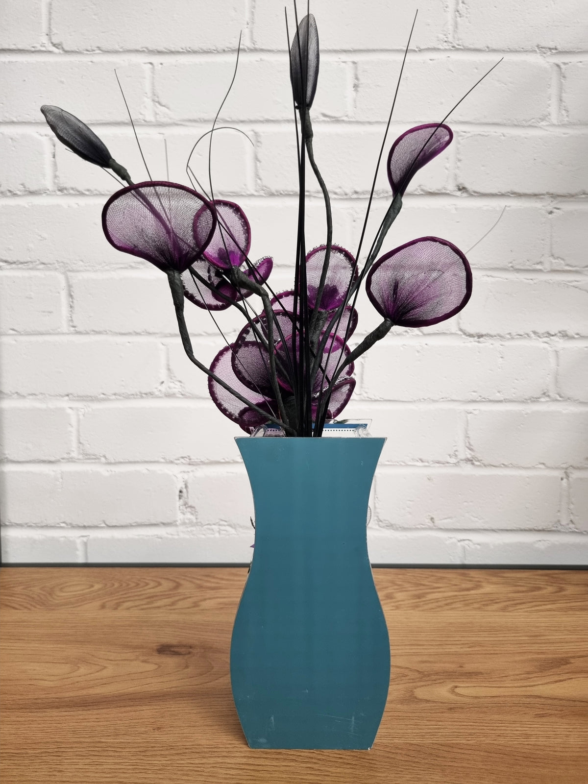 A decorative floral arrangement with purple mesh calla-lily-like blooms and dark stems in a teal-blue vase, set on a wooden surface against a white brick wall.