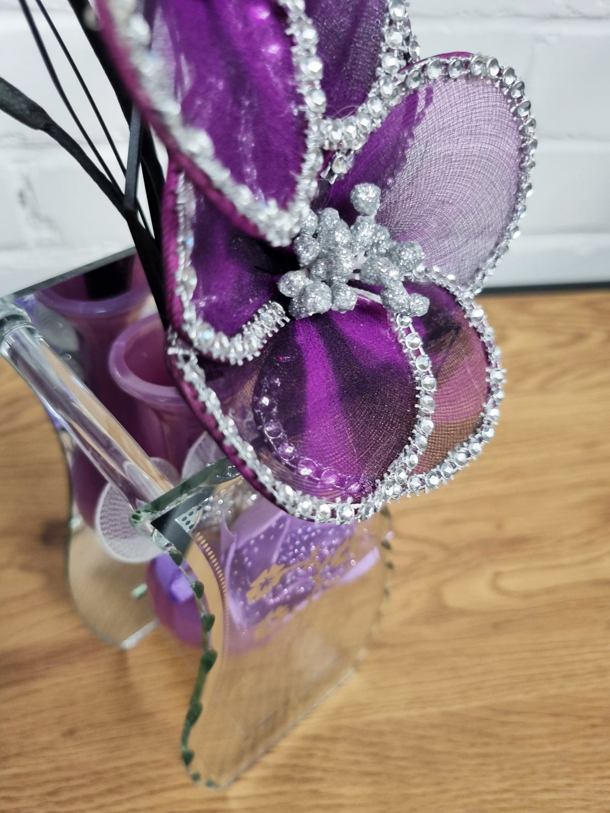 A close-up of a decorative purple fascinator or hair accessory with glittery purple mesh petals, silver rhinestone trim, and a silver floral center, held in a clear container on a wooden surface.