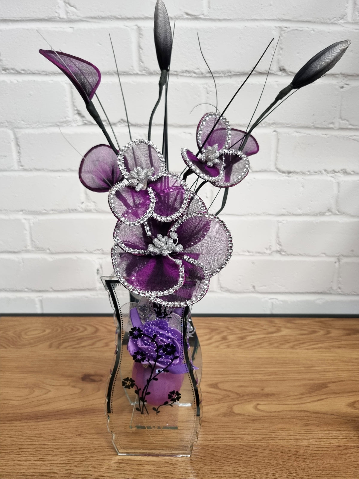 Purple artificial floral arrangement with rhinestone-embellished petals and long dark feather-like stems, displayed in a clear glass vase on a wooden surface against a white brick wall.
