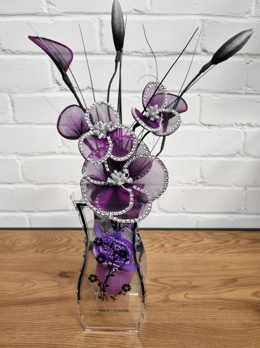 Purple artificial floral arrangement with rhinestone-embellished petals and long dark feather-like stems, displayed in a clear glass vase on a wooden surface against a white brick wall.