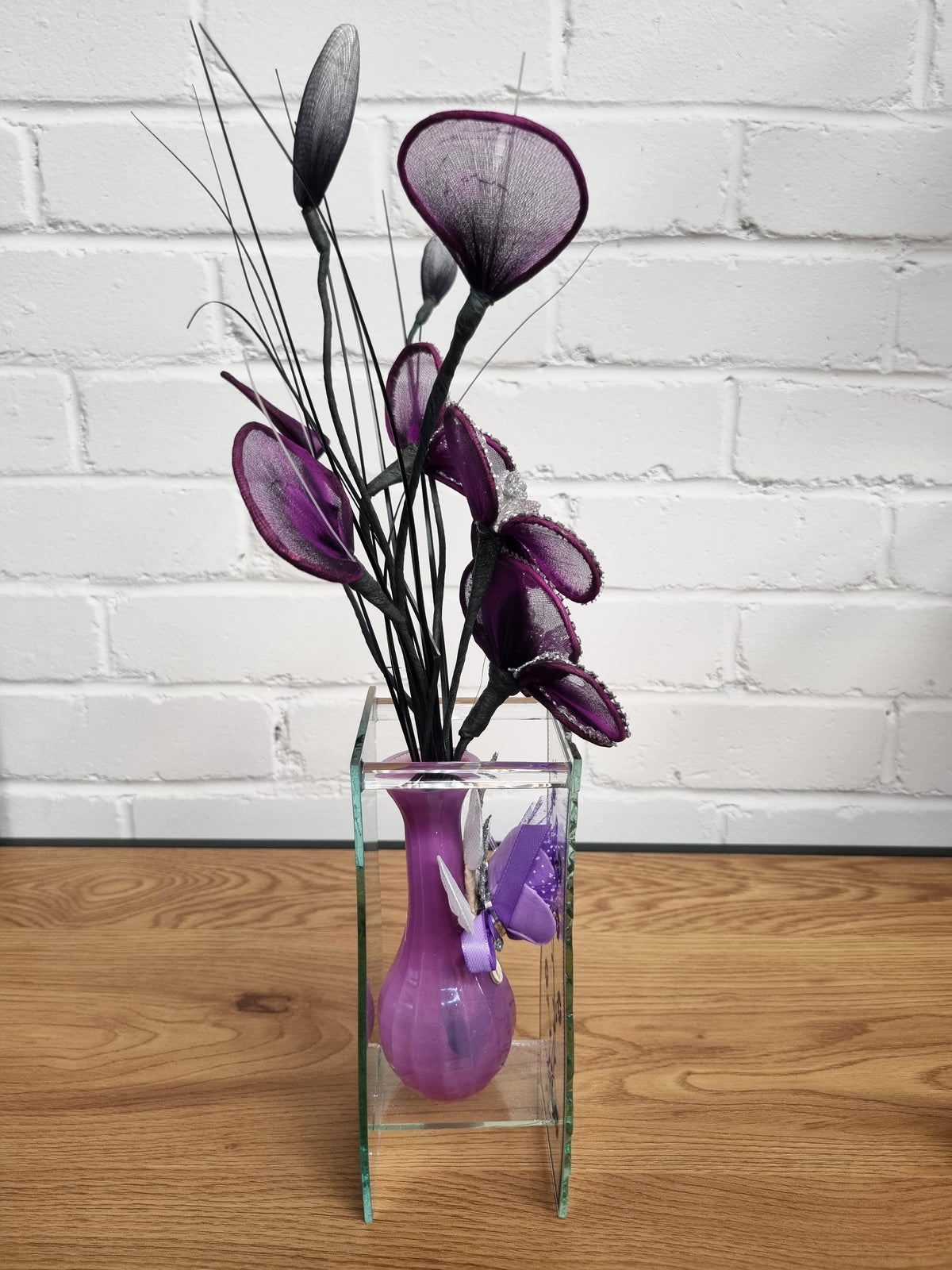 Purple artificial calla lilies with dark stems arranged in a clear square glass vase on a wooden tabletop against a white brick wall.