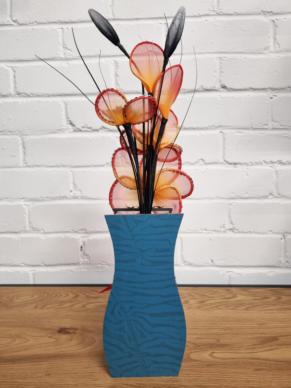 A decorative floral arrangement in a blue textured vase, featuring orange and peach translucent petal-like elements with black stems, set on a wooden floor against a white brick wall.