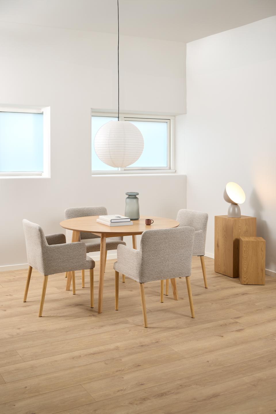A modern dining room setup featuring a round light oak wood table surrounded by four upholstered dining chairs in a light gray fabric. A white paper lantern pendant light hangs above the table, and a small wooden side table with a lamp sits to the right.