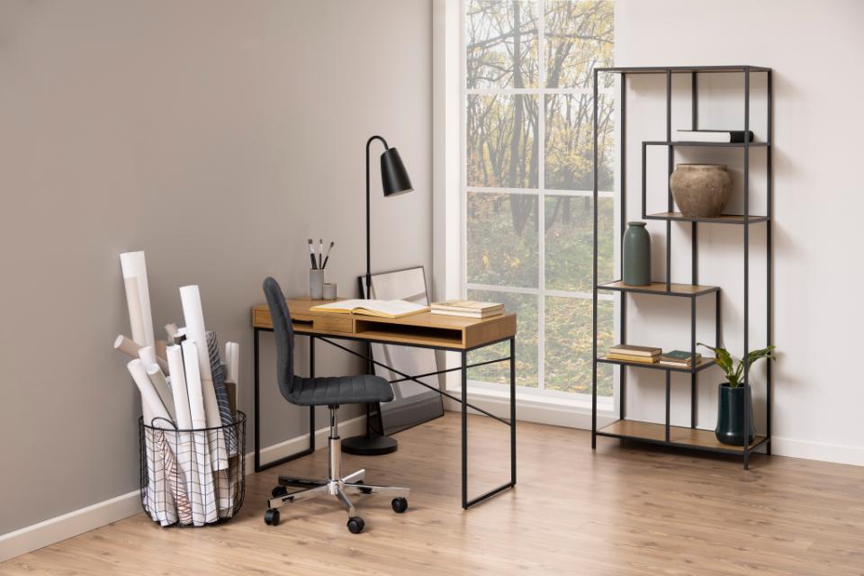 A modern home office setup featuring a wooden desk with a black metal frame, a black swivel office chair, a black gooseneck desk lamp, a wire basket organizer filled with rolled papers, and a tall black metal shelving unit decorated with vases and books, set against a neutral beige wall and wood floor.