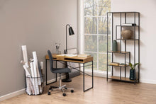 Load image into Gallery viewer, A modern home office setup featuring a wooden desk with a black metal frame, a black swivel office chair, a black gooseneck desk lamp, a wire basket organizer filled with rolled papers, and a tall black metal shelving unit decorated with vases and books, set against a neutral beige wall and wood floor.
