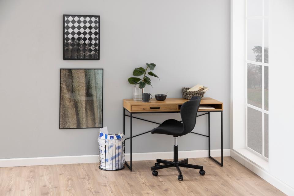 A modern home office setup featuring a wooden desk with a single drawer and black metal frame, paired with a black swivel office chair. The desk is styled with a small potted plant, a mug, and a basket, set against a light gray wall with framed art and a window on the right.