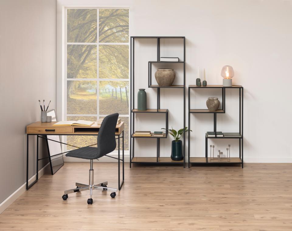 A modern home office setup featuring a light wood desk with a black metal frame, a black swivel office chair, and a matching black metal shelving unit with wood shelves. The room has light wood floors, beige walls, and a large window showing a tree view.