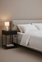 Load image into Gallery viewer, A modern bedroom scene featuring a black metal nightstand with a lit white table lamp, a small vase with a green sprig, and books on the shelf, positioned next to a bed with white bedding and a light gray upholstered headboard.
