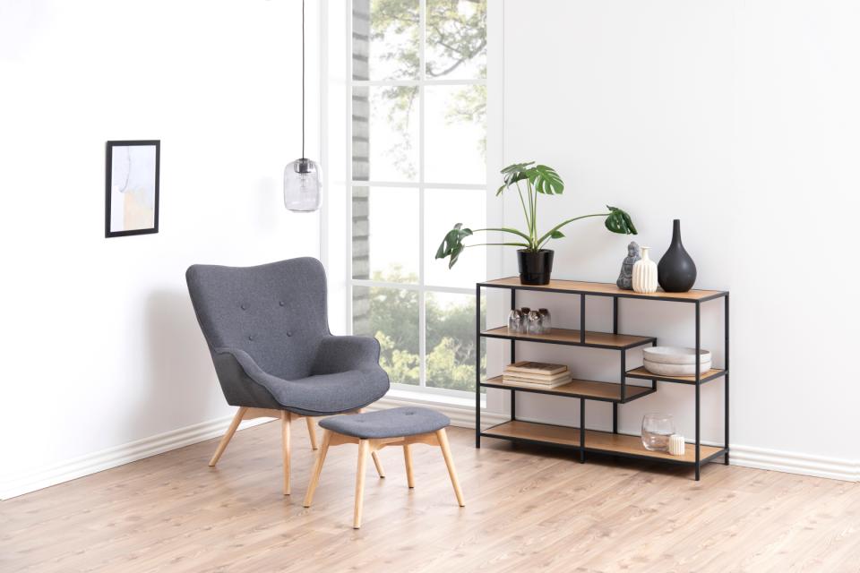 A modern gray upholstered armchair with a matching ottoman sits beside a black metal shelving unit with light wood shelves in a bright room with a window and wood floor.