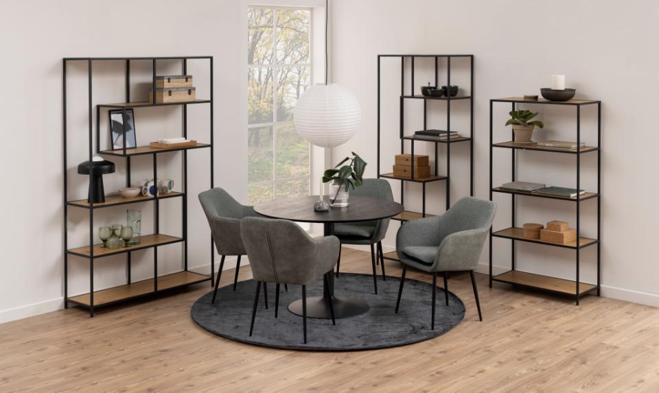 A styled interior scene showing a round dining table with four gray upholstered chairs on a round dark rug, surrounded by four black metal shelving units with wood shelves in a bright room with a window and wood floor.