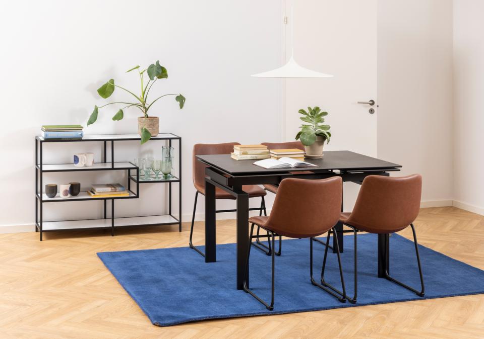 A modern dining setup in a bright room, featuring a black rectangular table with four brown upholstered chairs on a blue rug, alongside a black metal shelving unit with books and plants.