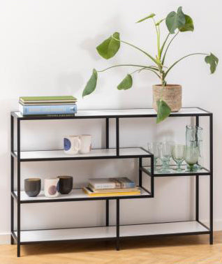 A modern black metal and glass shelving unit styled with books, mugs, glasses, and a potted plant, set against a plain white wall and light wood floor.