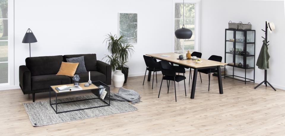 A modern living room and dining area featuring a black fabric sofa, a light wood coffee table with a black metal frame, a dining table set with black chairs, and a tall black shelving unit against a white wall.