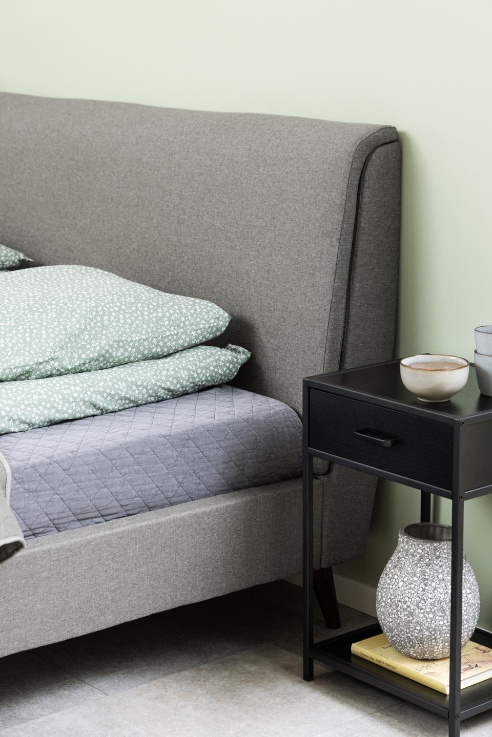 Gray upholstered bed with green bedding and a black nightstand with decorative items.
