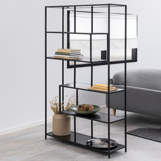 A modern black bookcase with five shelves, asymmetrically arranged, filled with books, plants, and decorative items, placed in a room with a grey sofa.