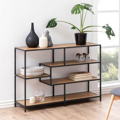 A Seaford stylish bookcase shelf unit featuring a combination of black metal frames and oak wood shelves, with dimensions 114x35x78 cm, displayed in a room setting with decorative items on top.