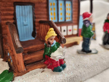 Load image into Gallery viewer, A miniature holiday village scene featuring a small log cabin with a blue door and windows, glittery white faux snow, and painted figurines of children. One child figurine in the foreground wears a yellow hat and green coat, sitting on the cabin steps.
