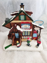 Load image into Gallery viewer, A decorative miniature village building set depicting a rustic ski school cabin with snow on the roof and base, small figures of children in winter clothing, and signage including 'Sugar Pine Ski School' and 'Snowboarda'.
