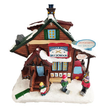 Load image into Gallery viewer, A small decorative village figurine of a winter ski school building with snow on the roof and ground, a red sign reading "Sugar Pine Ski School," a blue sign reading "Snowboarda Ski Les Bouç," and three tiny children standing in front on a snowy base.
