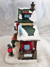 Load image into Gallery viewer, A small decorative winter village house figurine with snow-covered roofs, colorful windows, and tiny people near it, photographed on a soft white fabric background.
