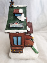 Load image into Gallery viewer, A small decorative house ornament with a green textured roof covered in white faux snow, brown wooden siding, blue window panes, and a sign reading "CLASSIC HOME OF J. PATRIOT.
