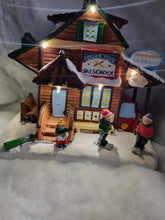 Load image into Gallery viewer, A decorative Christmas village figurine of a ski school cabin with snow-covered roofs, warm lights, and three small child characters standing in front on a snowy base.
