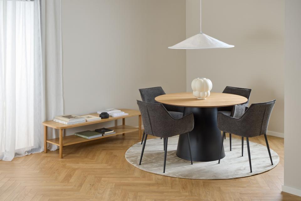A modern dining set featuring a round light oak wood Soli table with a black pedestal base, surrounded by four dark gray upholstered chairs on a round white rug. The scene is set in a minimalist room with light beige walls, a herringbone wood floor, a white pendant light, and a small wooden side table.
