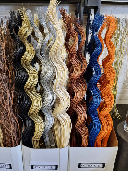 Multiple tall bundles of wavy decorative willow twig bunches displayed in white bins, shown in several colors including black, tan, cream, copper, blue, and orange. Bin labels read HOMESTREET.