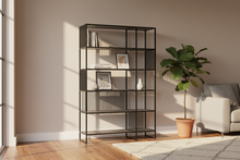 Load image into Gallery viewer, Black metal bookshelf in a room with a plant and sofa
