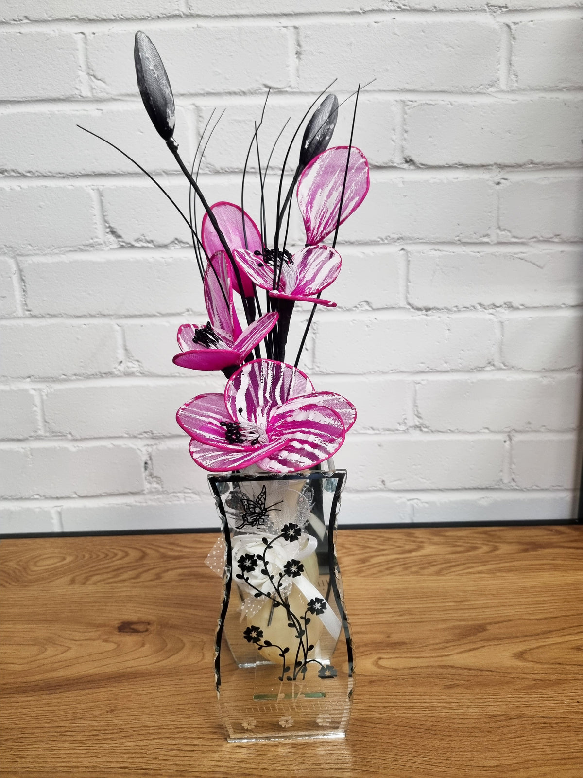 A decorative floral arrangement in a clear glass vase with black floral illustrations, set on a wooden surface against a white brick wall. The arrangement features vibrant pink and white striped flowers with black centers and long, thin black seed pods.