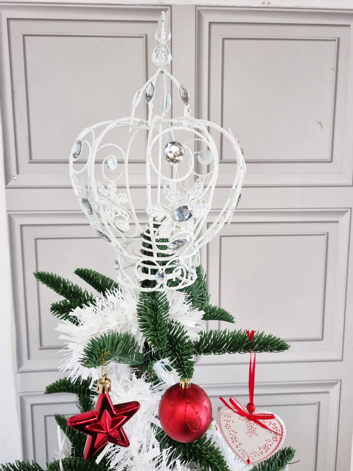 A white decorative metal tree topper with ornate scrollwork and hanging silver accents, displayed on a small green and white Christmas tree with red ornaments in front of a light gray paneled door.