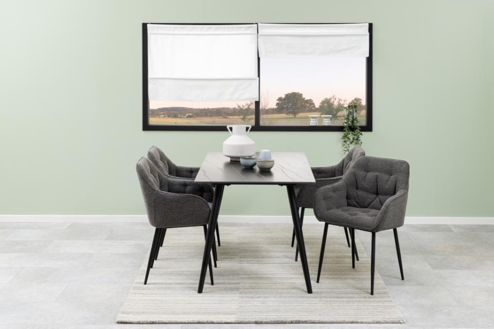 A modern dining set featuring a rectangular dark gray stone table and four tufted gray upholstered chairs, arranged on a light gray rug in front of a window with a sage green wall.