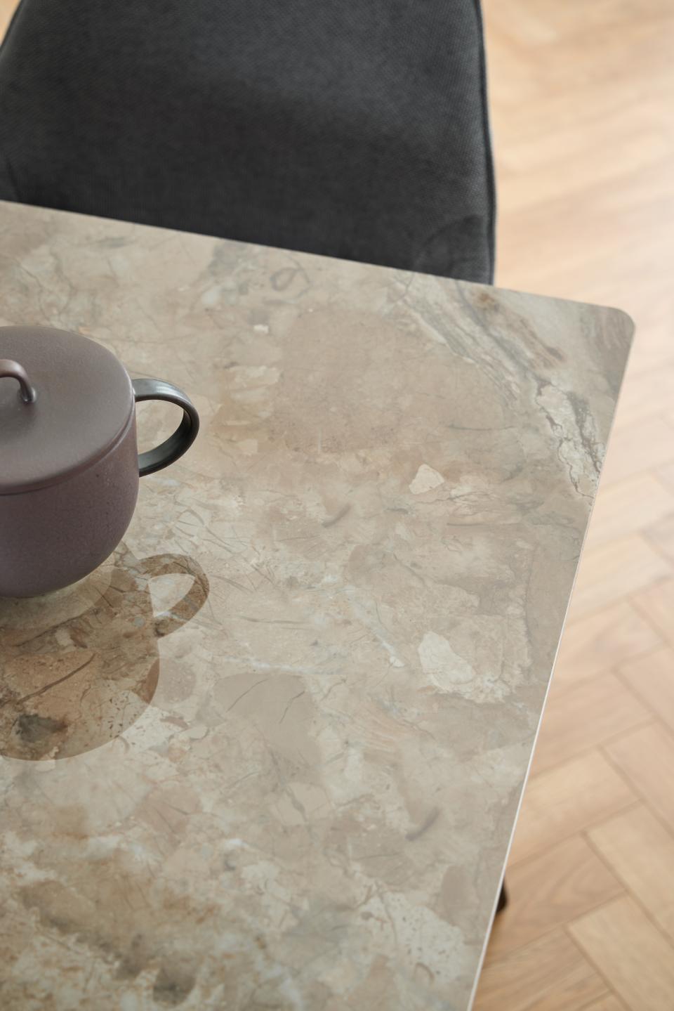 A close-up of a rectangular table with a beige and gray marble-look surface, with part of a dark upholstered chair visible at the top and a small teapot on the left. Light wood flooring is visible on the right.