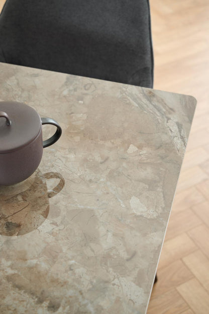 A close-up of a rectangular table with a beige and gray marble-look surface, with part of a dark upholstered chair visible at the top and a small teapot on the left. Light wood flooring is visible on the right.