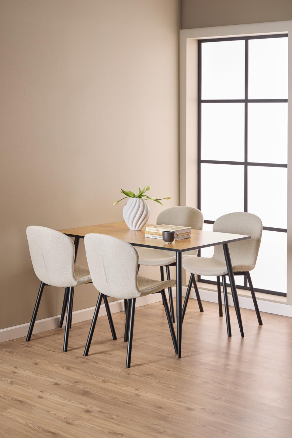 A modern dining set featuring a rectangular light wood table with a black frame and four upholstered dining chairs in a warm beige color with black legs, set in a minimalist dining room with beige walls and light wood flooring.