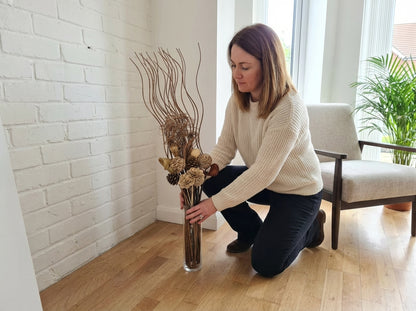 A woman kneels on a light wood floor next to a white brick wall, holding a tall clear glass vase filled with dried branches, pinecones, and dried flowers.