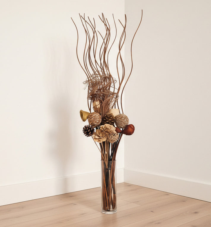 A tall, decorative dried floral arrangement in a clear cylindrical glass vase, placed on a light wood floor against a plain off-white wall corner.