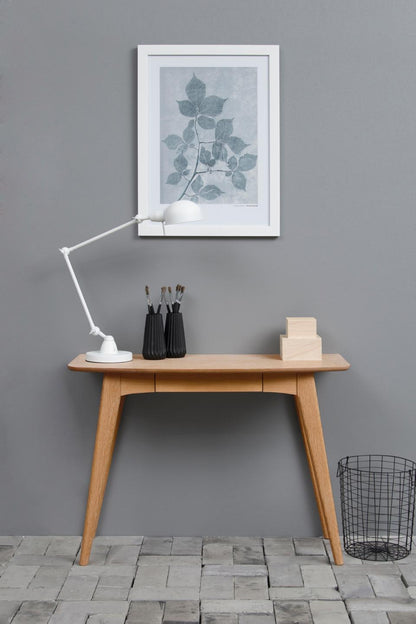 Wooden desk with lamp, plant artwork, and small items against a gray wall.