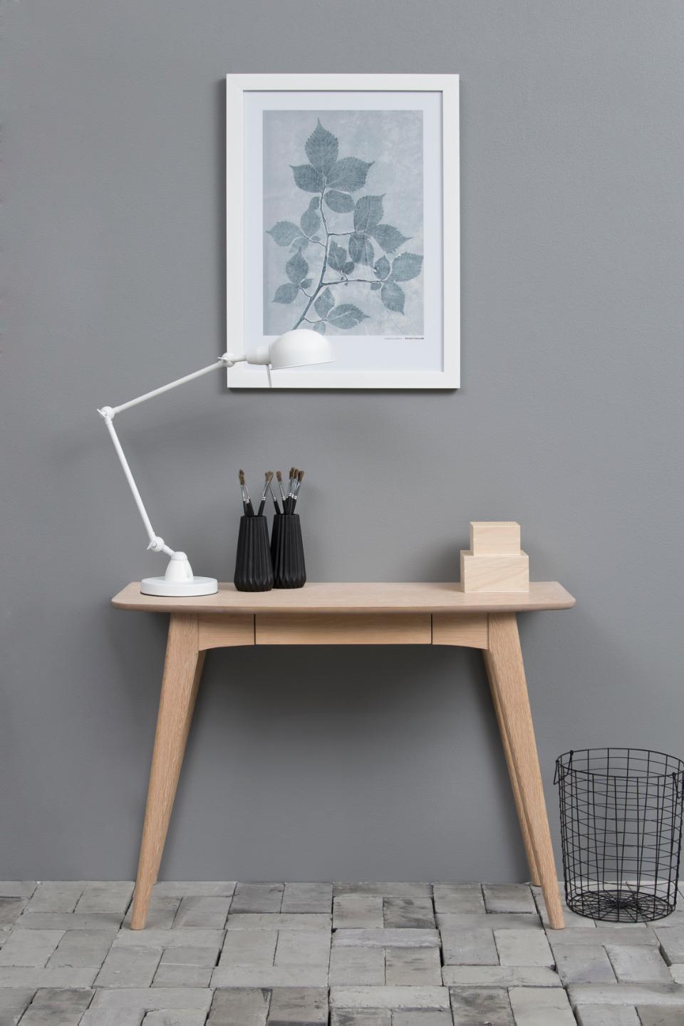 Wooden desk with lamp, plant artwork, and small items against a gray wall.