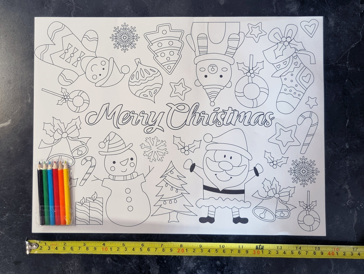 A black and white Christmas coloring page featuring Santa Claus, a snowman, stockings, ornaments, candy canes, and the words 'Merry Christmas,' shown on a dark textured surface with a yellow tape measure and colored pencils underneath for scale.