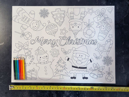 A black and white Christmas coloring page featuring Santa Claus, a snowman, stockings, ornaments, candy canes, and the words 'Merry Christmas,' shown on a dark textured surface with a yellow tape measure and colored pencils underneath for scale.