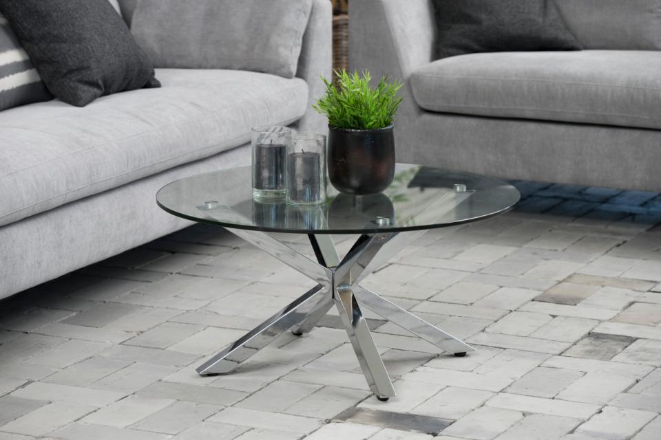 heaven clear glass table with a silver metal cross base on a stone floor next to grey sofas with a plant and candles on top