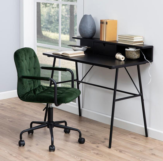 A black rectangular office desk with a drawer and a lower shelf, featuring metal legs. A green chair is also visible in the background, suggesting the desk is set in a home environment.