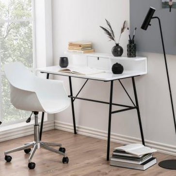 A modern home office setup featuring a white desk with black metal legs, a white swivel chair, a black floor lamp, and a gray corkboard on the wall, set in a bright room with light wood floors and a window.